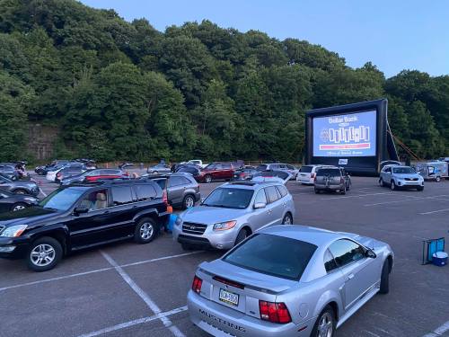 A drive-in movie setup with cars parked in a lot, featuring a large inflatable screen and trees in the background.