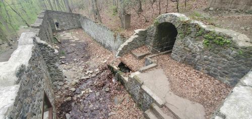 Ruins of a stone structure in a forest, with a small stream and a person walking in the background among fallen leaves.