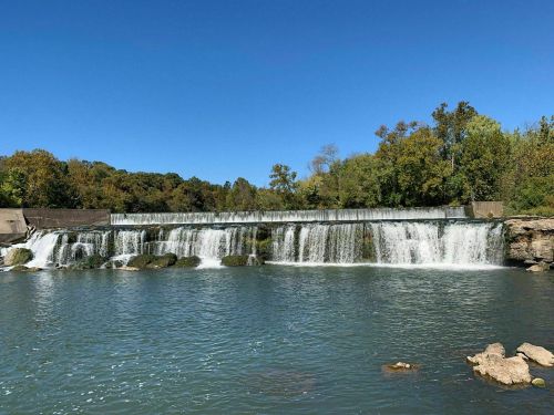A serene waterfall cascading over a dam into a calm river, surrounded by lush green trees under a clear blue sky.