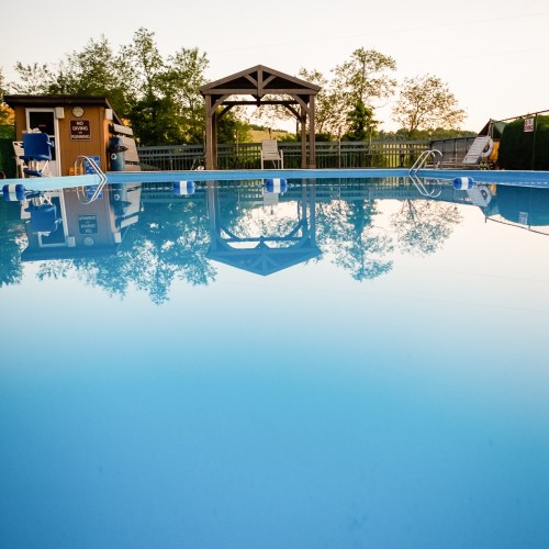 A serene swimming pool reflecting a gazebo and trees, surrounded by a wooden fence under a clear sky.