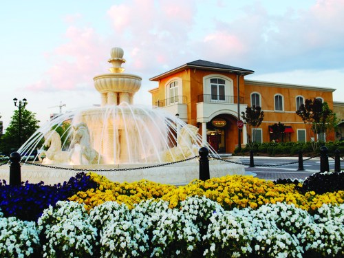 A decorative fountain with flowing water, surrounded by colorful flowers, in front of a building under a pastel sky.