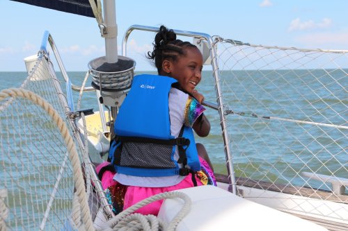 A smiling girl in a life jacket sits on a sailboat, enjoying the view of the water on a sunny day.