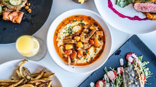 A vibrant dish of creamy grits topped with vegetables and herbs, surrounded by various plates of food and a drink.