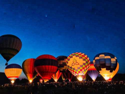 Colorful hot air balloons illuminated against a twilight sky, with a crowd gathered below.