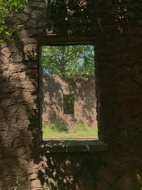 A stone wall with a window frame, revealing another stone wall and greenery outside. Sunlight filters through.