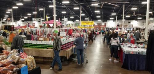 A bustling indoor market with various vendor booths, shoppers browsing, and colorful displays of goods.