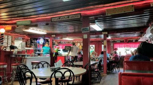 A colorful diner interior with red accents, tables, and patrons enjoying their meals. Neon signs and decor add charm.