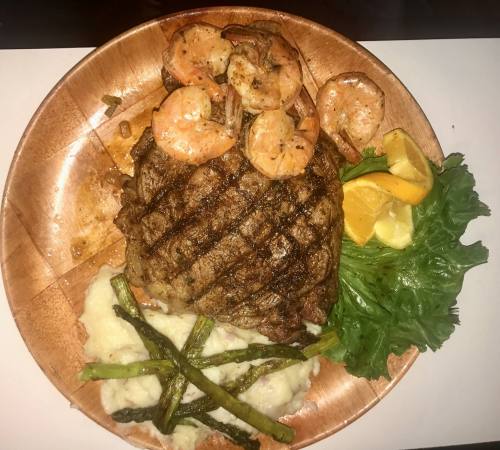 A grilled steak topped with shrimp, served with mashed potatoes, asparagus, and lemon wedges on a wooden plate.