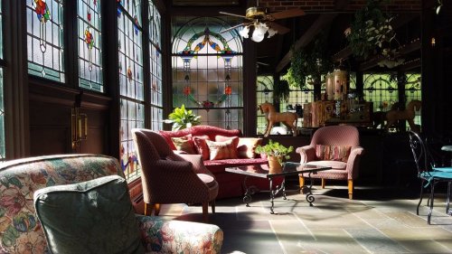 Cozy seating area with stained glass windows, plants, and vintage decor in a sunlit room.