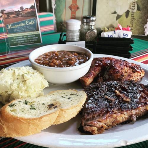 A plate of barbecue featuring ribs, brisket, baked beans, coleslaw, and two slices of bread on a colorful table setting.