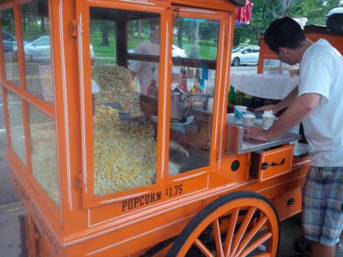 A vendor prepares popcorn at an orange cart filled with popcorn, with a price sign of $1.75 visible.