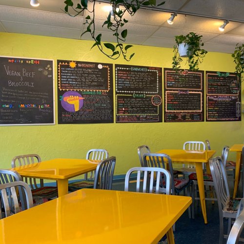 Brightly colored café interior with yellow tables, metal chairs, and chalkboard menus on the walls. Green plants hang above.