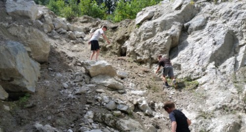 Three children climbing a rocky hillside surrounded by greenery on a sunny day.