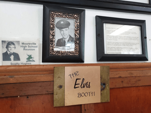 A wall display featuring a photo of a soldier, a reunion sign, and a plaque labeled "The Elvis Booth."