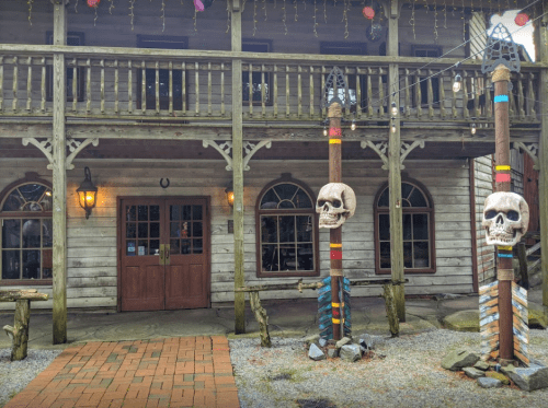 A rustic wooden building with skull decorations on totem poles and warm lighting, set in a gravel area.