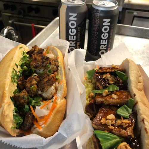 Two gourmet sandwiches on a tray: one with spiced shrimp and veggies, the other with grilled meat and toppings, plus two cans of soda.
