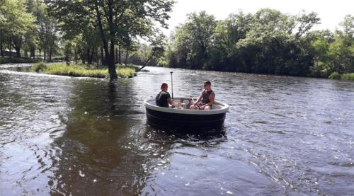 Two people sit in a small boat on a calm river, surrounded by trees and greenery.