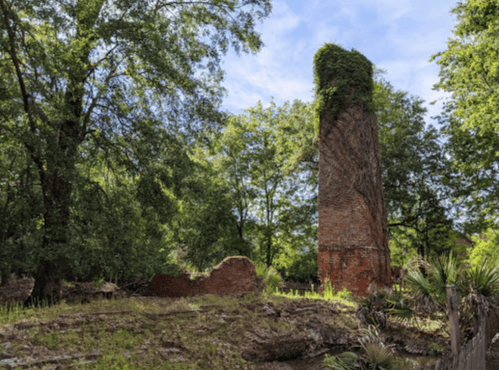 A tall, ivy-covered brick chimney stands among trees, with remnants of a stone structure nearby.