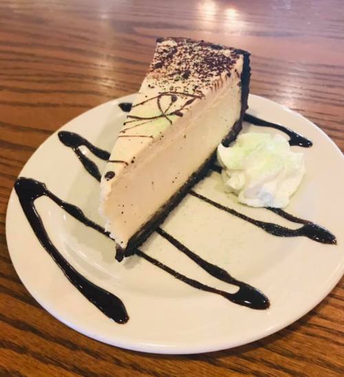 A slice of creamy cheesecake on a plate, drizzled with chocolate sauce and topped with whipped cream.
