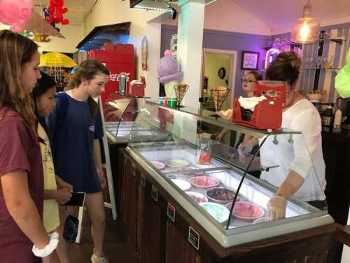 Three girls look at a variety of ice cream flavors in a shop, while a staff member serves ice cream behind the counter.