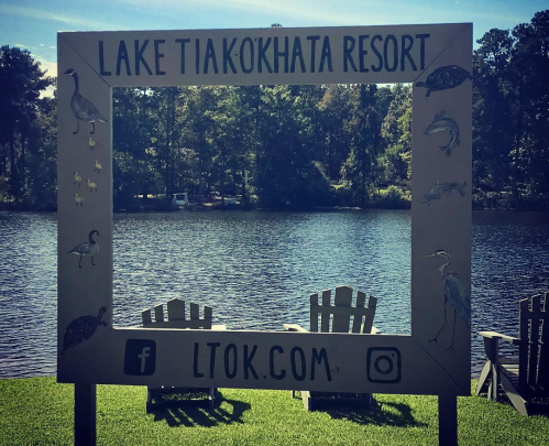 A sign for Lake Tiak-Okhata Resort frames a scenic view of the lake with chairs in front.
