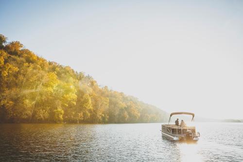 A boat glides on a serene lake surrounded by autumn-colored trees under a bright, sunny sky.