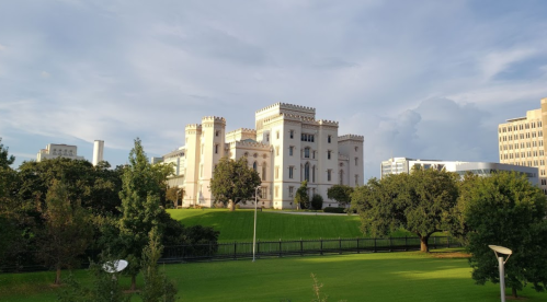 Historic castle-like building surrounded by green lawns and trees under a partly cloudy sky.