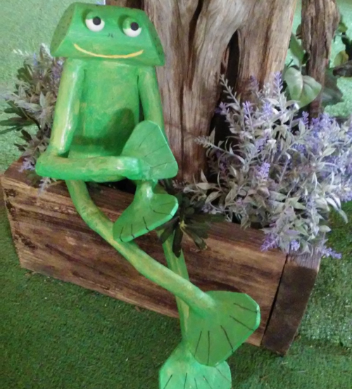 A whimsical green frog sculpture sitting on a wooden box, surrounded by purple flowers and greenery.