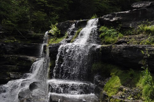 A serene waterfall cascading over rocky ledges, surrounded by lush greenery and sunlight filtering through the trees.