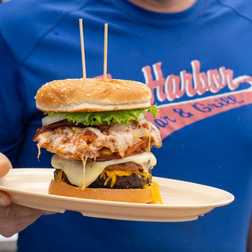 A towering burger with layers of cheese, lettuce, and chicken, held on a plate by a person in a blue shirt.