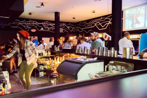 A lively bar scene with patrons socializing, a bartender mixing drinks, and a large screen displaying a video.