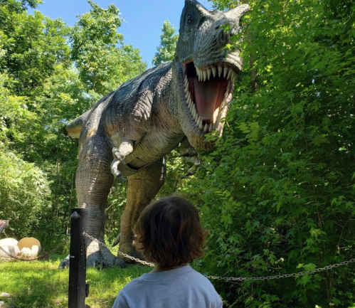 A child stands in front of a roaring dinosaur statue surrounded by lush greenery.