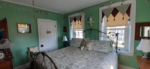 Cozy bedroom with green walls, a floral bedspread, and decorative windows featuring patterned shades.