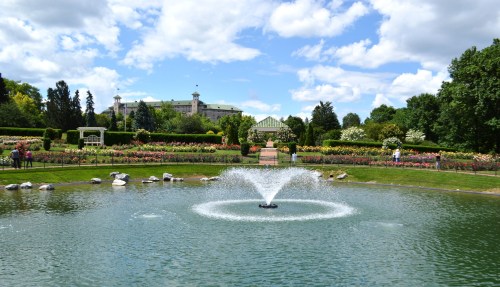 A serene garden with a fountain in the foreground, surrounded by colorful flowers and lush greenery under a blue sky.