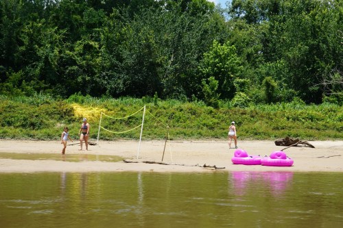 Three people on a sandy beach near a river, with a volleyball net and pink inflatable floats nearby. Lush greenery in the background.