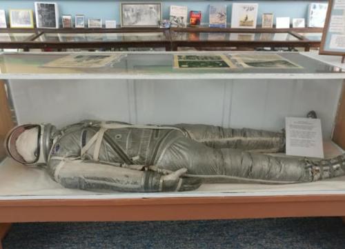 A display case containing a vintage astronaut suit lying flat, with informational materials in the background.