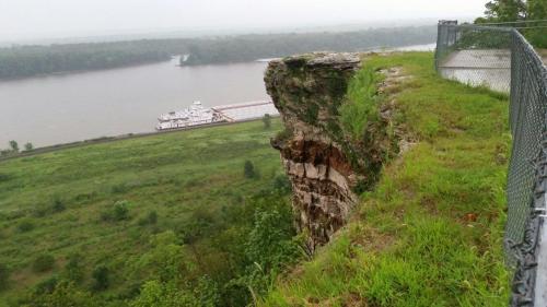 A rocky cliff overlooks a river with a barge, surrounded by lush greenery and a fence along the edge.