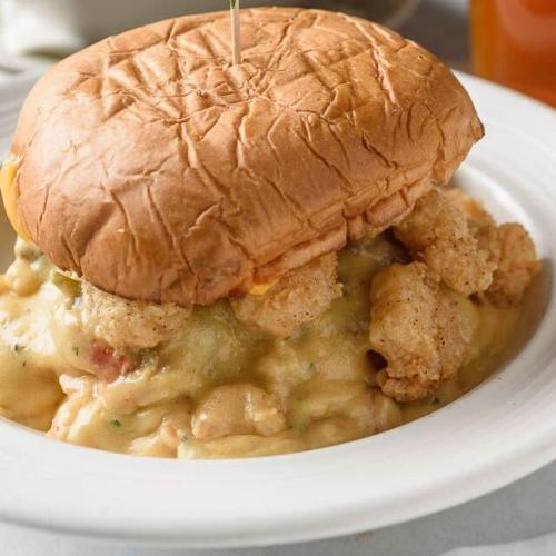 A sandwich with a toasted bun sits atop creamy macaroni and cheese, topped with crispy fried shrimp.