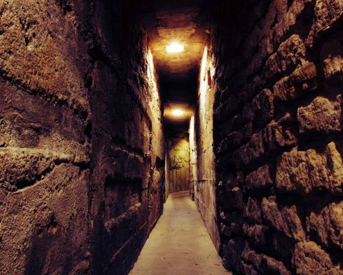 A dimly lit corridor with stone walls and ceiling lights, creating a mysterious and atmospheric setting.