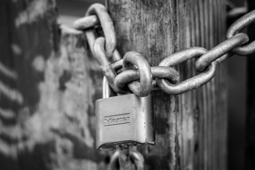 A close-up of a silver padlock secured with a chain on a weathered wooden fence, in black and white.