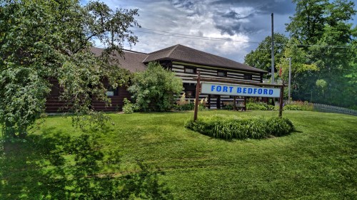 A log cabin building with a sign reading "Fort Bedford," surrounded by greenery and under a cloudy sky.