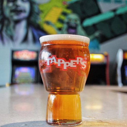 A glass of amber beer with a frothy head, featuring the logo "TAPPER'S," set against a colorful arcade backdrop.