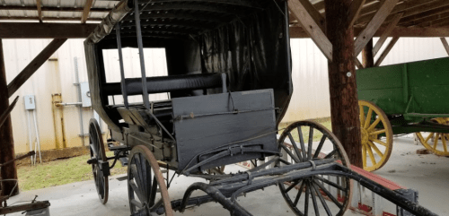 A vintage horse-drawn carriage with a black canopy, displayed under a wooden shelter.