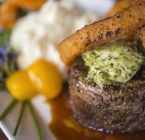 A gourmet steak topped with herb butter, served with mashed potatoes and garnished with vegetables.