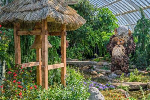 A whimsical garden scene featuring a large, furry creature carrying a rock, surrounded by lush greenery and flowers.