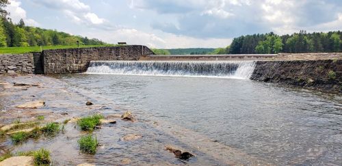 A serene view of a stone dam with water cascading over it, surrounded by lush greenery and a cloudy sky.