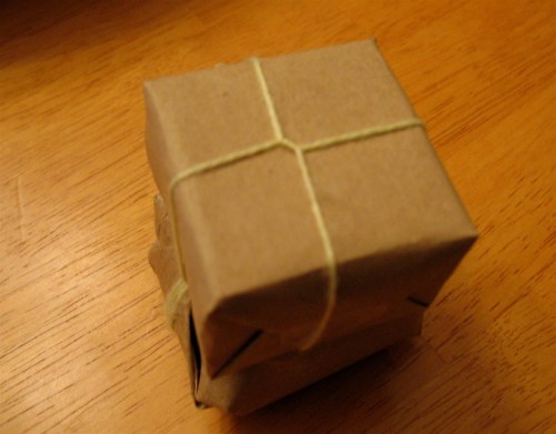 A small, wrapped package made of brown paper, tied with a thin string, resting on a wooden surface.
