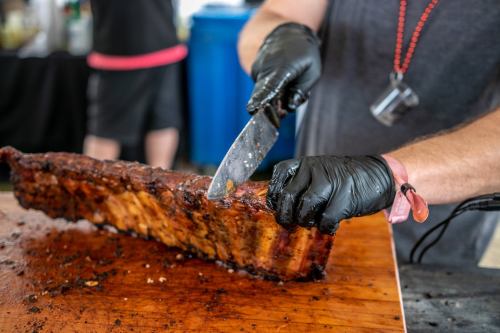 A person in black gloves slices a rack of ribs on a wooden cutting board.