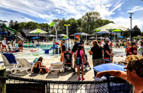 A busy water park scene with families, children, and colorful umbrellas around a pool on a sunny day.