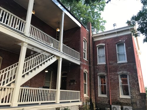 Historic brick building with a white staircase and balcony, surrounded by trees in a serene setting.